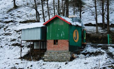 A wooden cabin blanketed in snow at Sissu village in Lahaul Valley, surrounded by Himalayan mountains